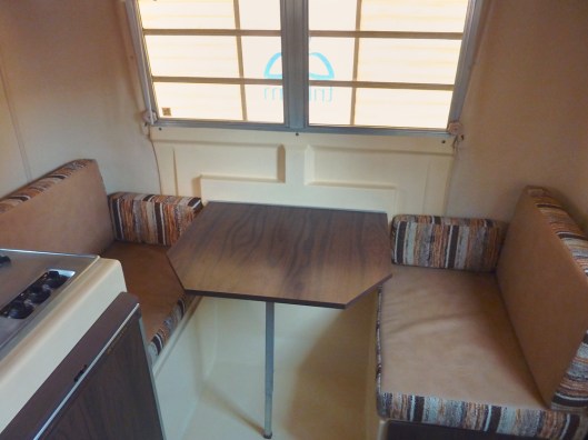 Front Dinette with vinyl side of the cushions on top, mini-blinds pulled up. The exterior fiberglass rock shield/window awning is filtering the light.